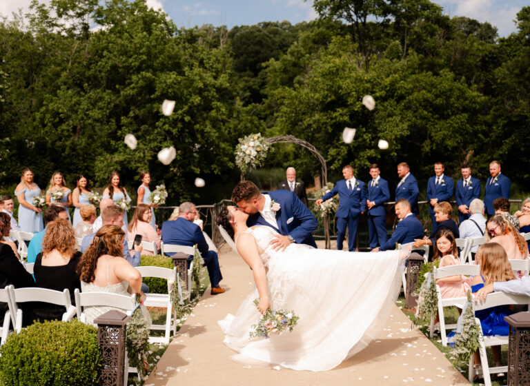 Bride and groom kiss at outdoor wedding, surrounded by guests tossing hats, with lush greenery in background.