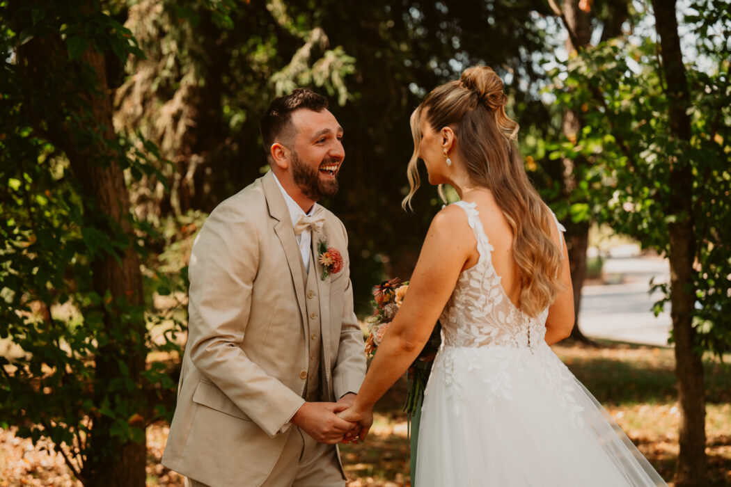 A bride and groom smiling and holding hands outdoors beneath green trees.