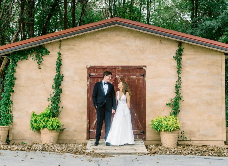 A bride and groom stand in wedding attire in front of a vine-covered building, gazing at each other.