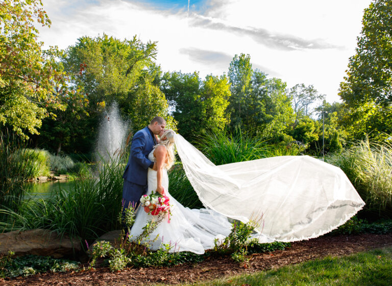 A bride and groom share a kiss near a pond, with her veil flowing and greenery surrounding them.